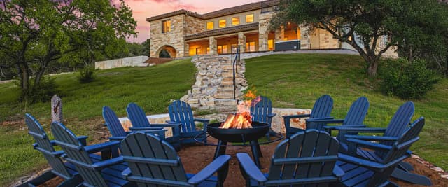 Blue Adirondack chairs arranged around a fire pit at sunset, with stone house and oak trees in background