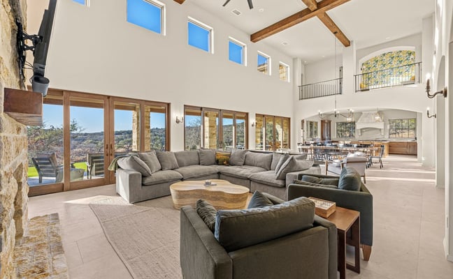 Cozy living room with stone fireplace, burning fire, armchair with blanket, books and mug on table, large windows overlooking landscape.